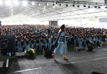 Woman in academic regalia crosses stage before large audience under enormous tent